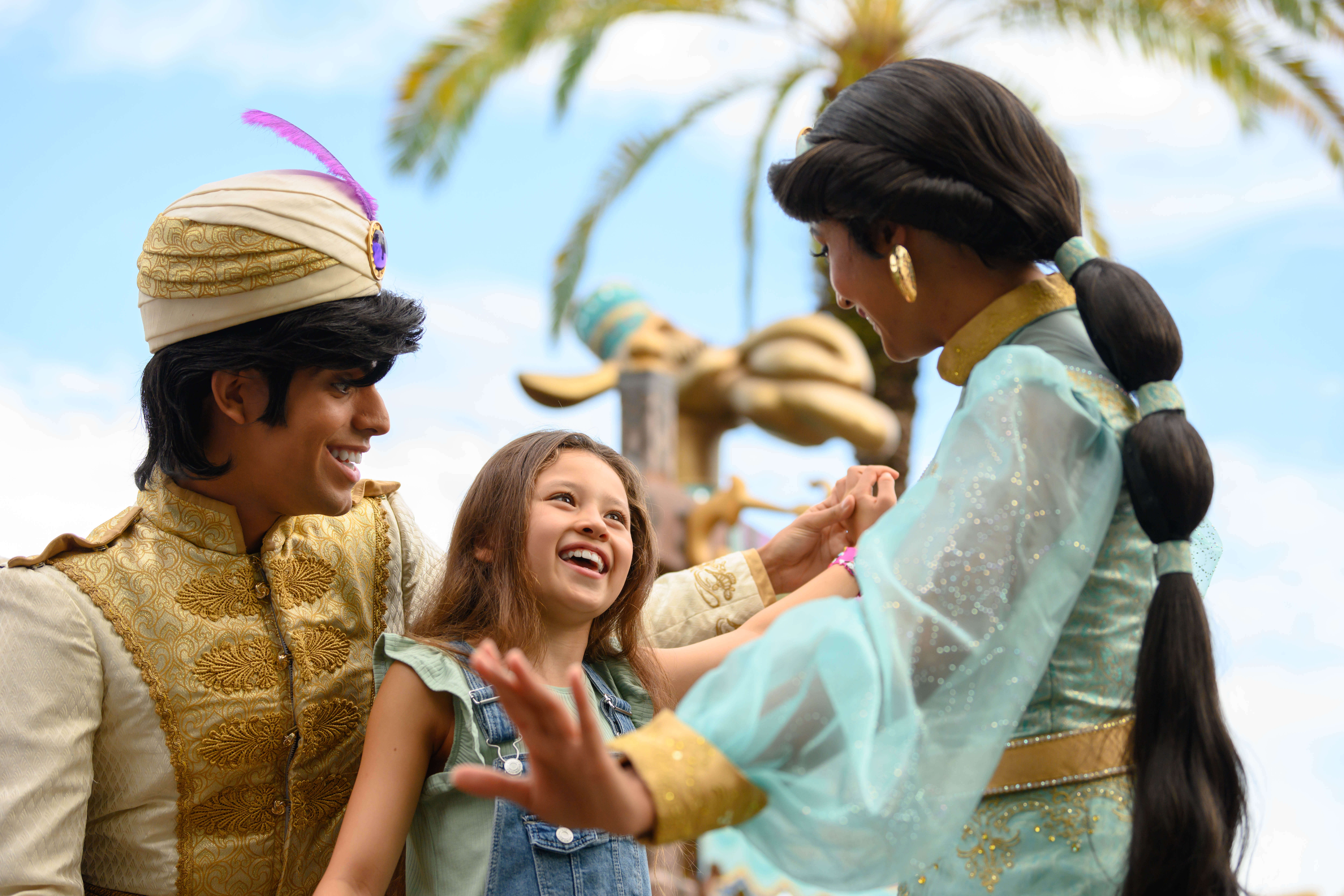 A smiling young girl happily holds hands with two people dressed as Aladdin and Jasmine in a bright outdoor setting with palm trees in the background.