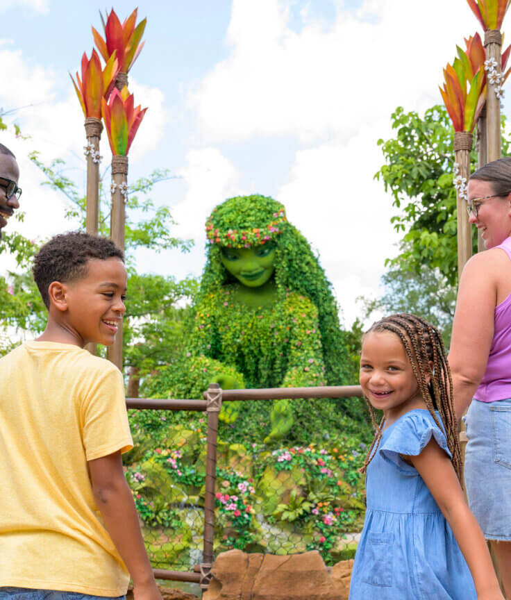 A smiling family of four stands in front of a large, green plant sculpture of a woman surrounded by tropical plants and flowers, enjoying a sunny day at a theme park.