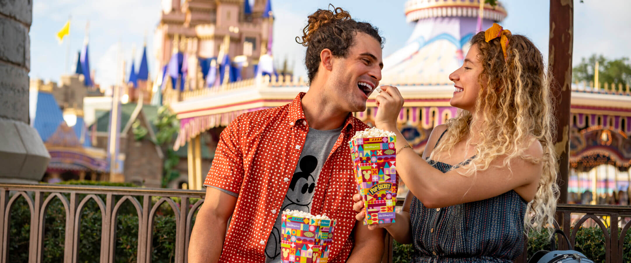 A smiling couple sits on a bench at a theme park, with a colorful castle in the background. The woman feeds popcorn to the man as they enjoy their snacks together. Both appear happy and playful.