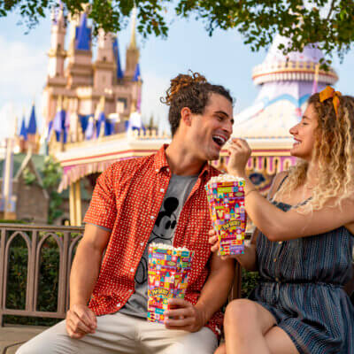 زوجان مبتسمان يجلسان على مقعد في متنزه ترفيهي، وفي الخلفية قلعة ملونة. تطعم المرأة الفشار للرجل بينما يستمتعان بوجباتهما الخفيفة معًا. يبدو كلاهما سعيداً ومرحاً.