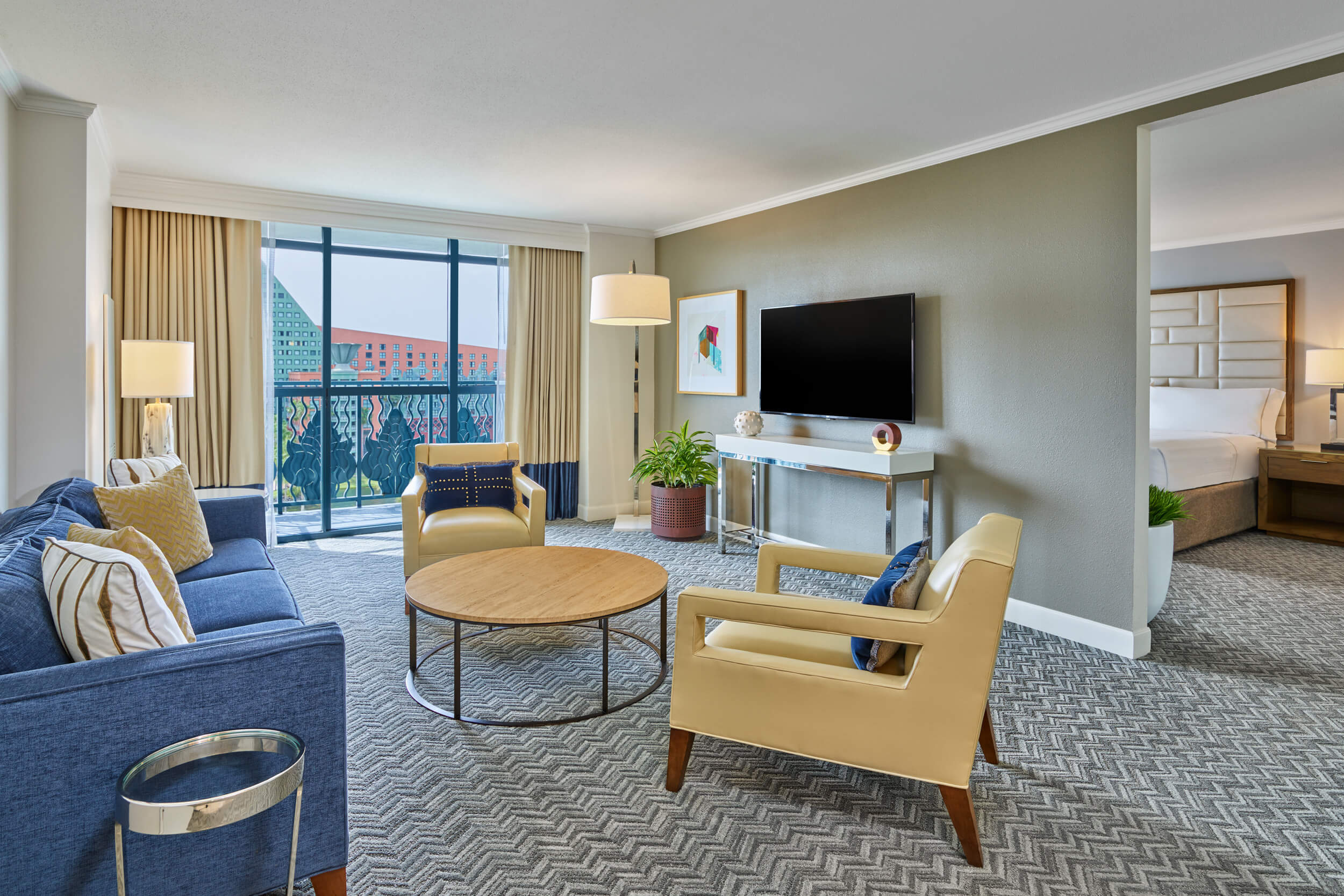 Modern hotel suite with a seating area featuring a blue sofa, two tan chairs, a round coffee table, TV on the wall, large window with balcony, and a partial view of a bedroom with a white bed.