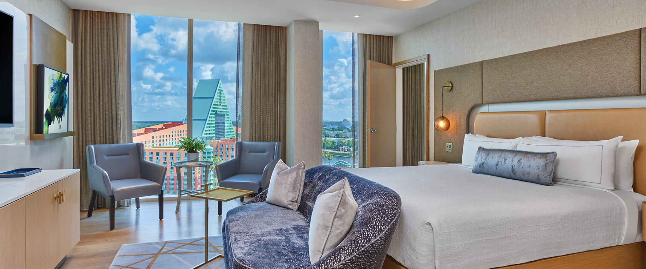 Modern hotel room with a large bed, a curved sofa, two chairs by a window, and a view of city buildings and a blue sky. The room has wood floors, neutral colors, and contemporary decor.