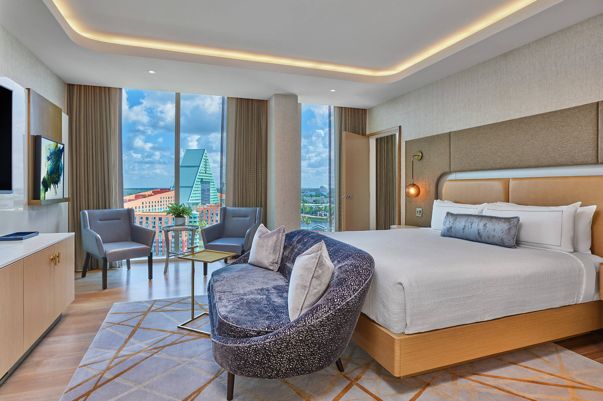 Modern hotel room with a large bed, a curved sofa, two chairs by a window, and a view of city buildings and a blue sky. The room has wood floors, neutral colors, and contemporary decor.