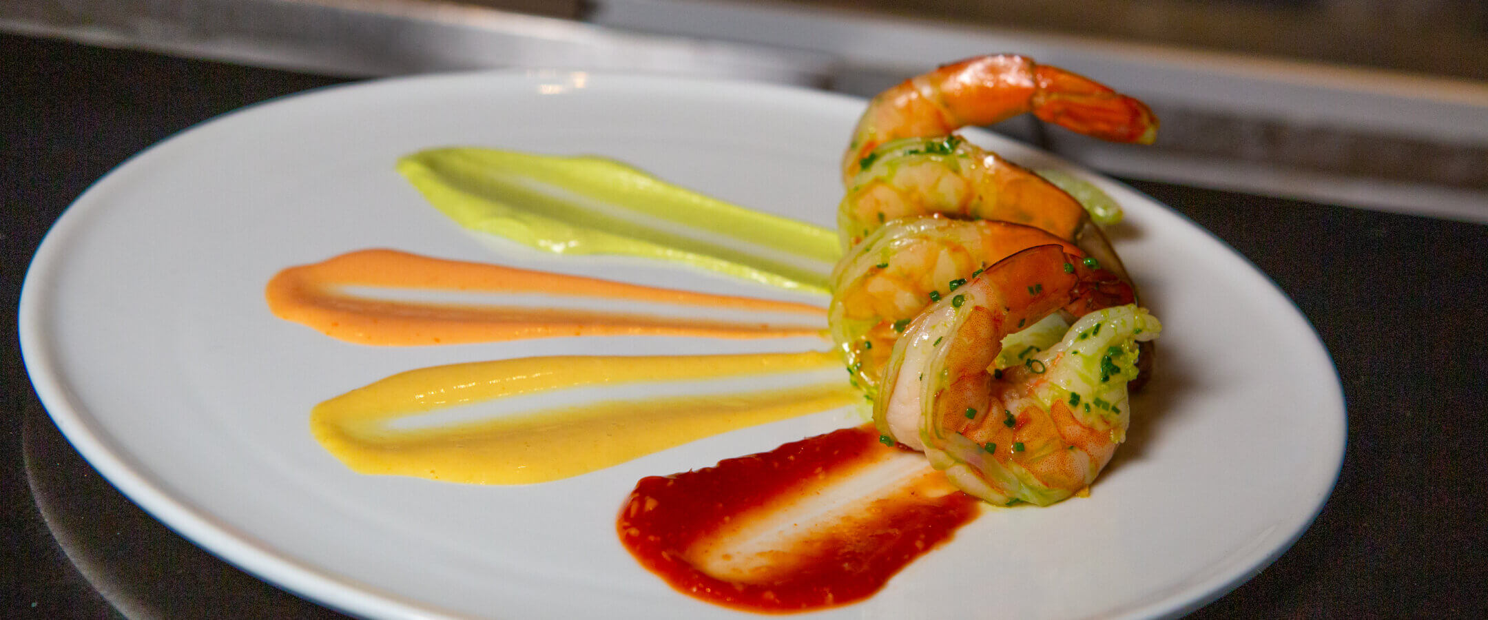 Three cooked shrimp are arranged on a white plate with colorful sauces—red, yellow, orange, and green—neatly brushed in parallel lines beside them, creating an artistic presentation.