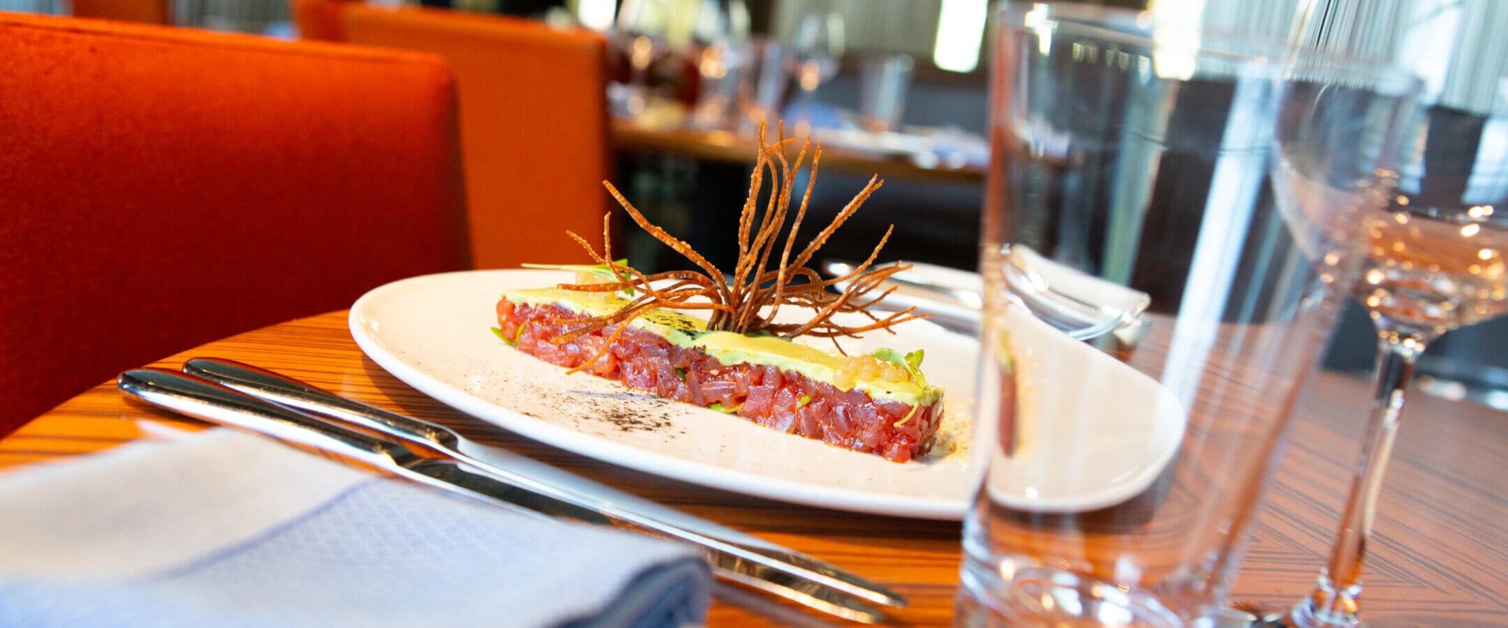 A close-up of a plated gourmet dish on a wooden restaurant table set with glassware, cutlery, and a blue napkin; orange chairs and a softly lit dining area are visible in the background.