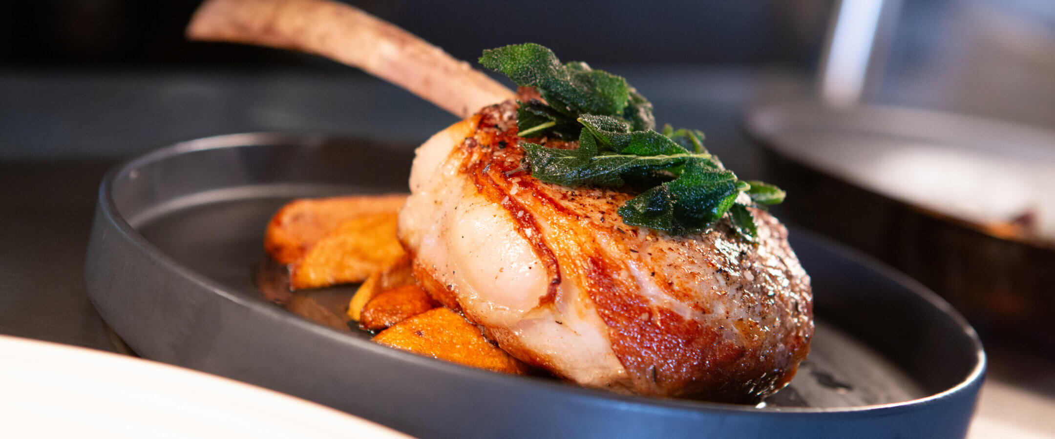 A cooked bone-in pork chop garnished with fresh herbs sits on top of roasted potato wedges on a dark plate. The dish is well-lit and served in a modern restaurant setting.