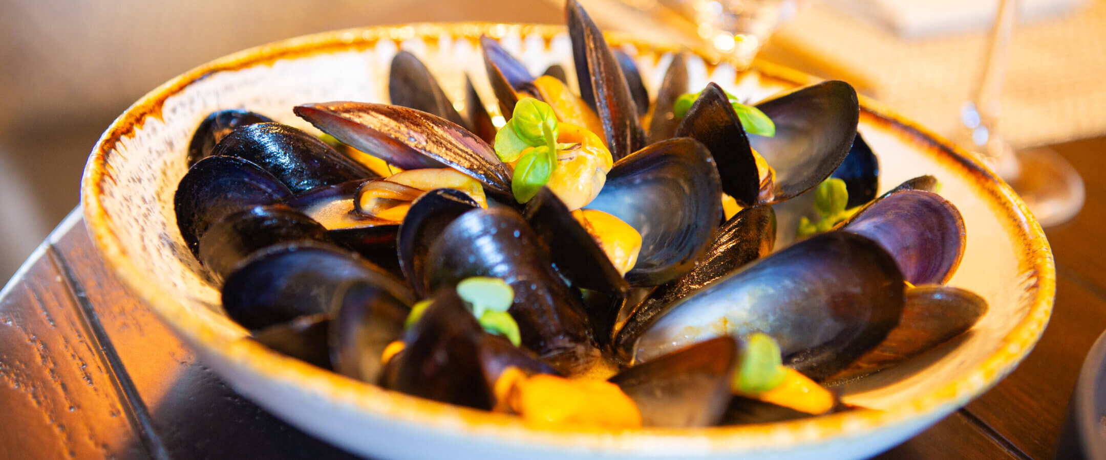 A bowl of steamed mussels garnished with fresh herbs, served on a ceramic plate on a wooden table, with wine glasses and napkins in the blurred background.