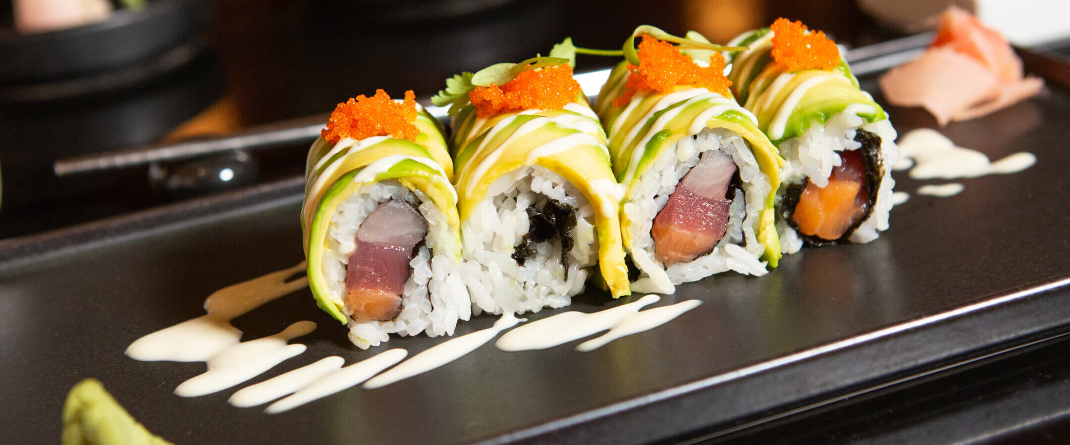 Four sushi rolls topped with avocado slices, orange roe, and drizzled with white sauce are arranged on a black plate, garnished with wasabi and pickled ginger, with chopsticks and other dishes in the background.