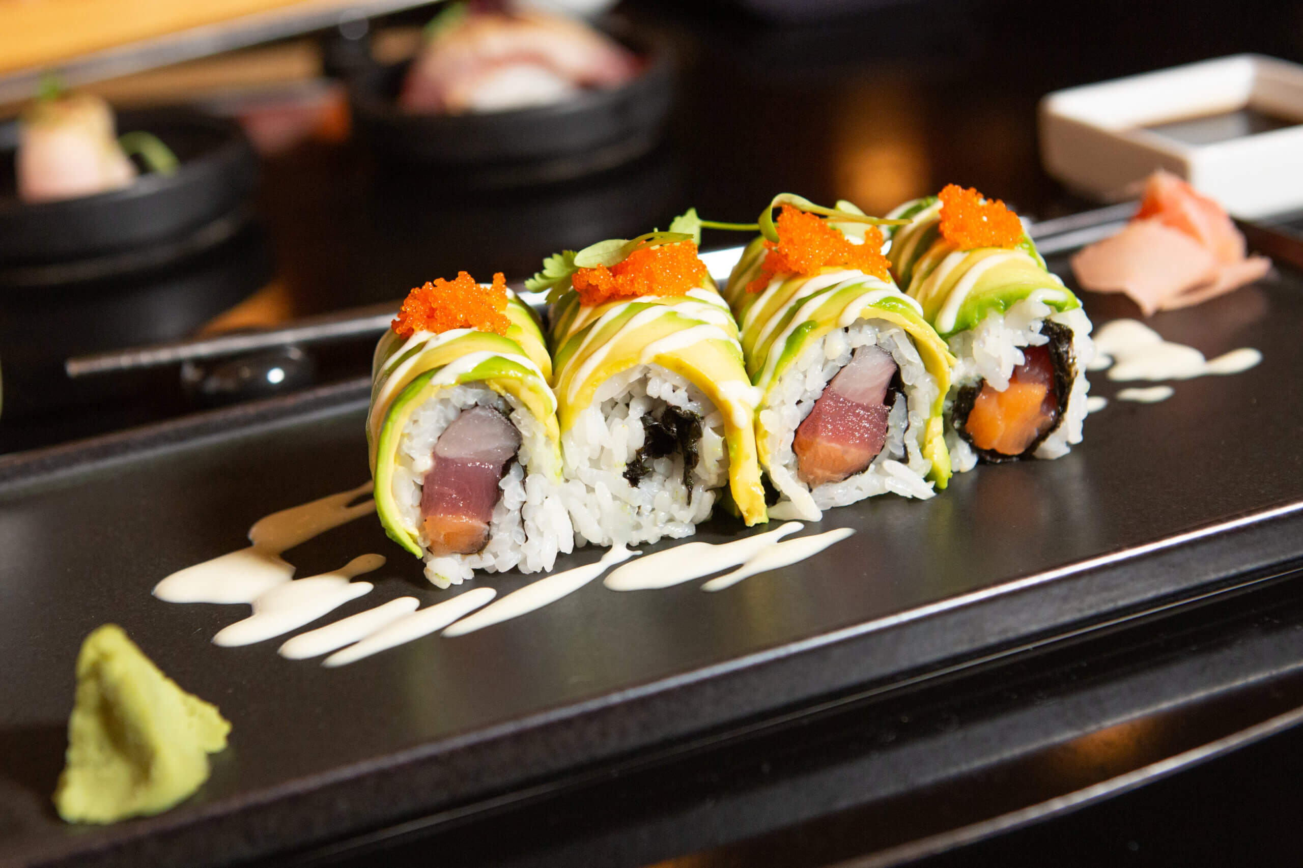 Four sushi rolls topped with avocado slices, orange roe, and drizzled with white sauce are arranged on a black plate, garnished with wasabi and pickled ginger, with chopsticks and other dishes in the background.