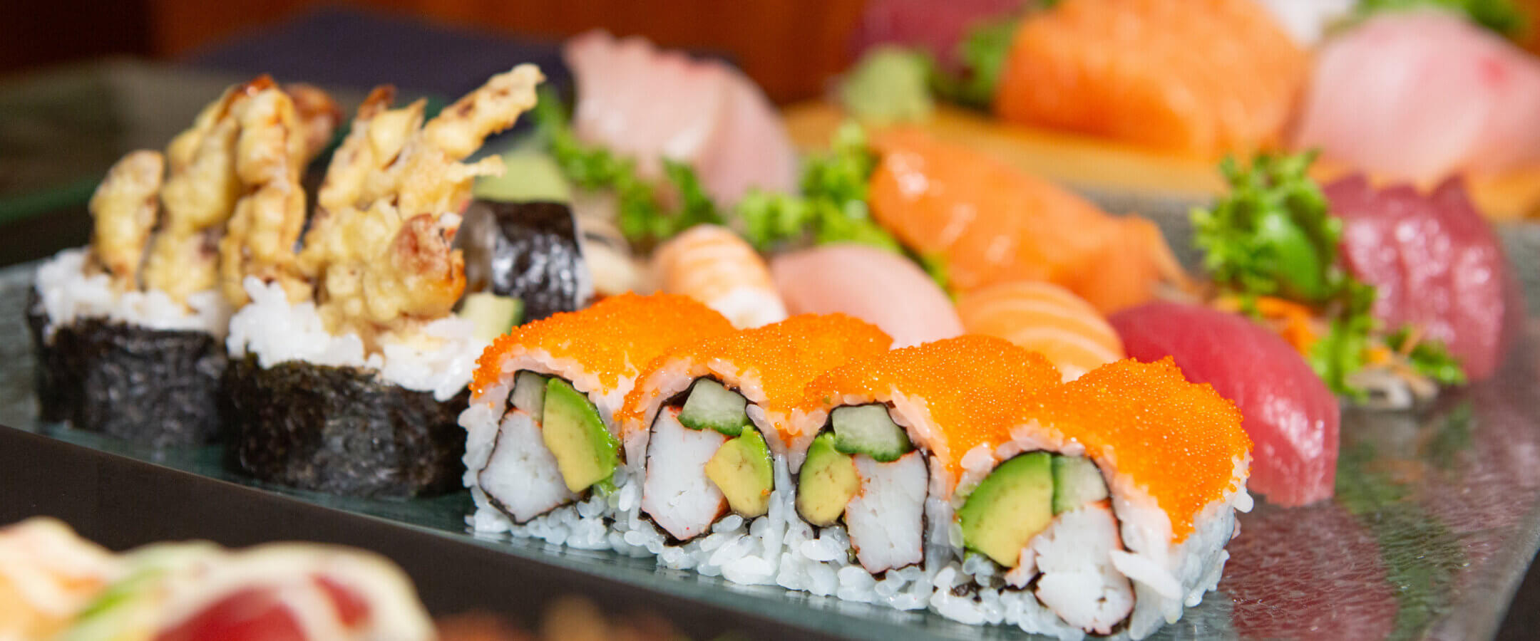 A close-up of assorted sushi rolls and sashimi on a platter, featuring sushi rolls with orange fish roe, avocado, and crab, along with pieces of nigiri and sashimi garnished with greens.