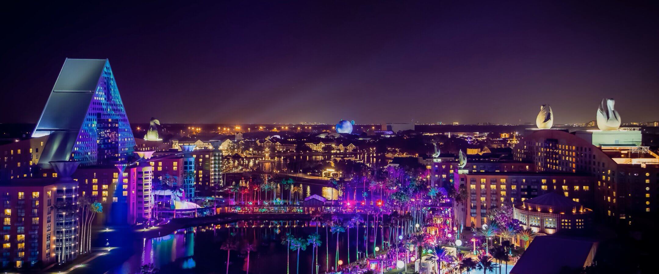 A vibrant, colorful night view of a cityscape featuring tall, uniquely shaped buildings, illuminated palm trees, and glowing purple and blue lights reflecting on water.