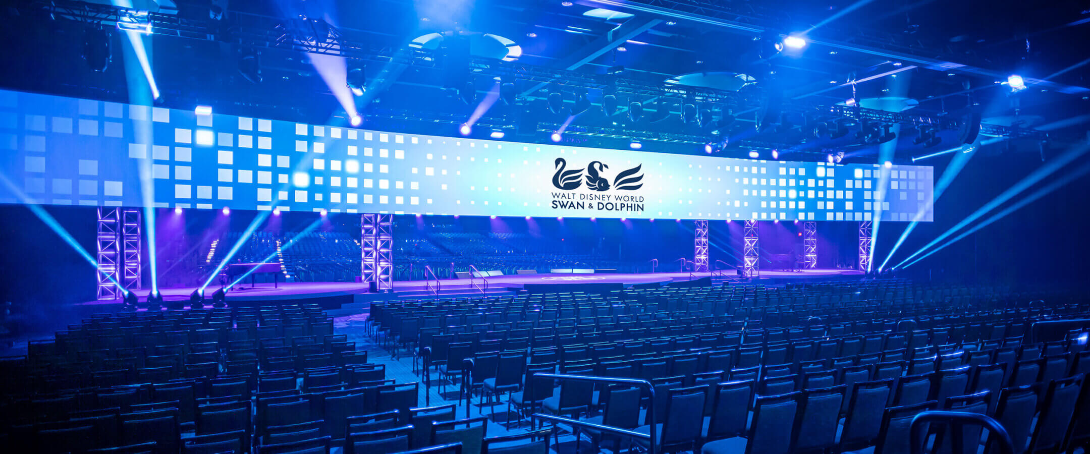 A large, modern event hall with rows of empty chairs faces a wide, illuminated stage featuring blue lighting and a digital screen displaying the Swan & Dolphin logo.