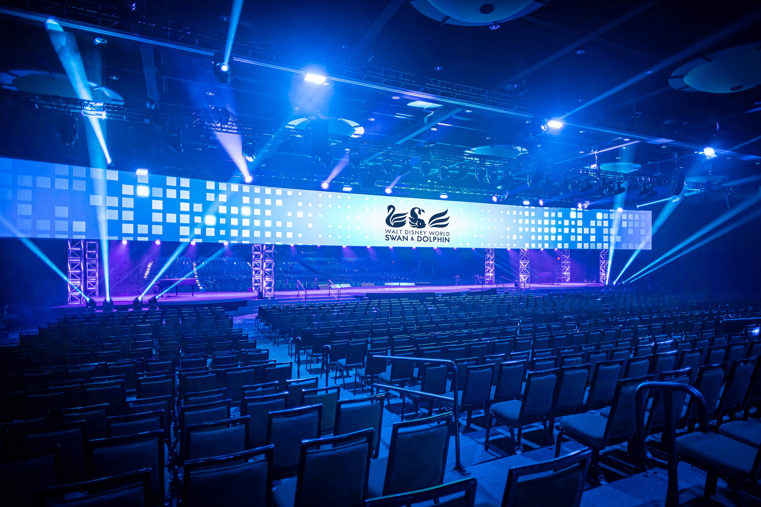 A large, modern event hall with rows of empty chairs faces a wide, illuminated stage featuring blue lighting and a digital screen displaying the Swan & Dolphin logo.