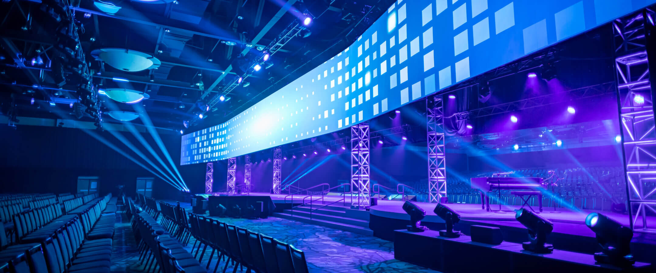 Large, modern event stage with bright blue and purple lighting, digital screen displaying pixel patterns, empty rows of chairs, and spotlights illuminating the spacious venue.