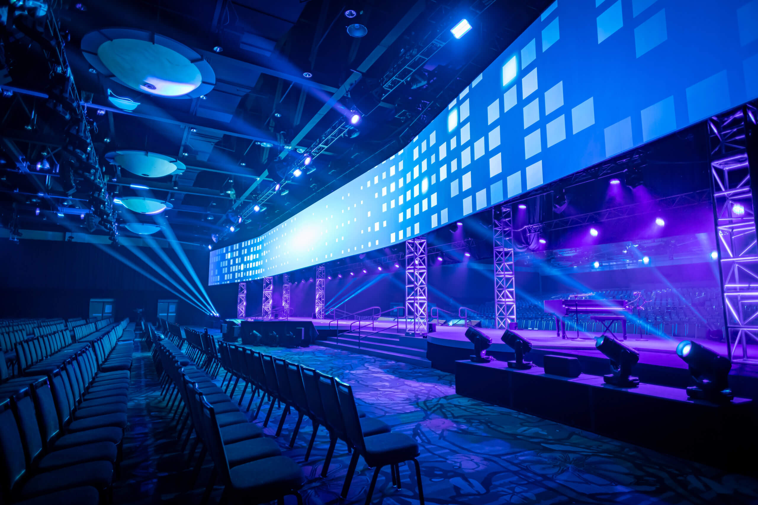 Large, modern event stage with bright blue and purple lighting, digital screen displaying pixel patterns, empty rows of chairs, and spotlights illuminating the spacious venue.