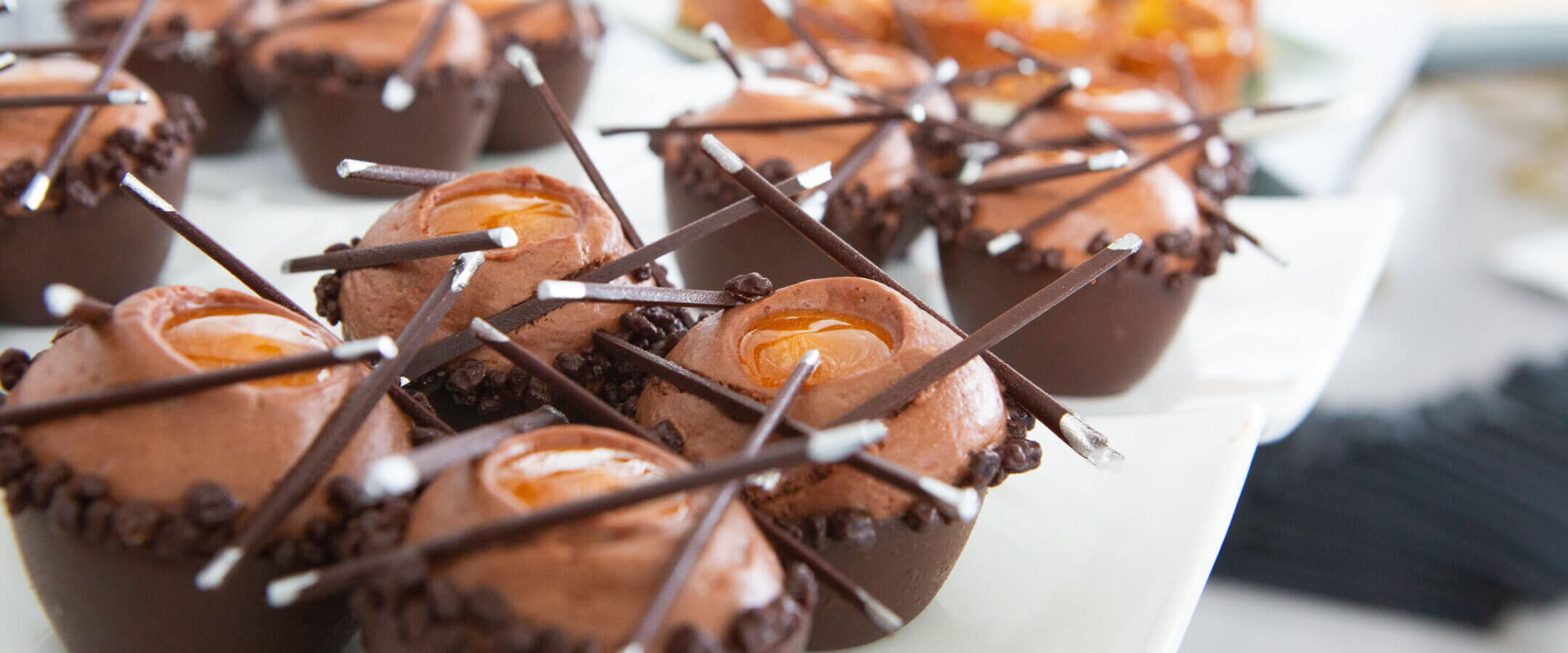 Chocolate desserts topped with swirls of chocolate mousse, caramel centers, and thin chocolate sticks, arranged neatly on a white serving platter.