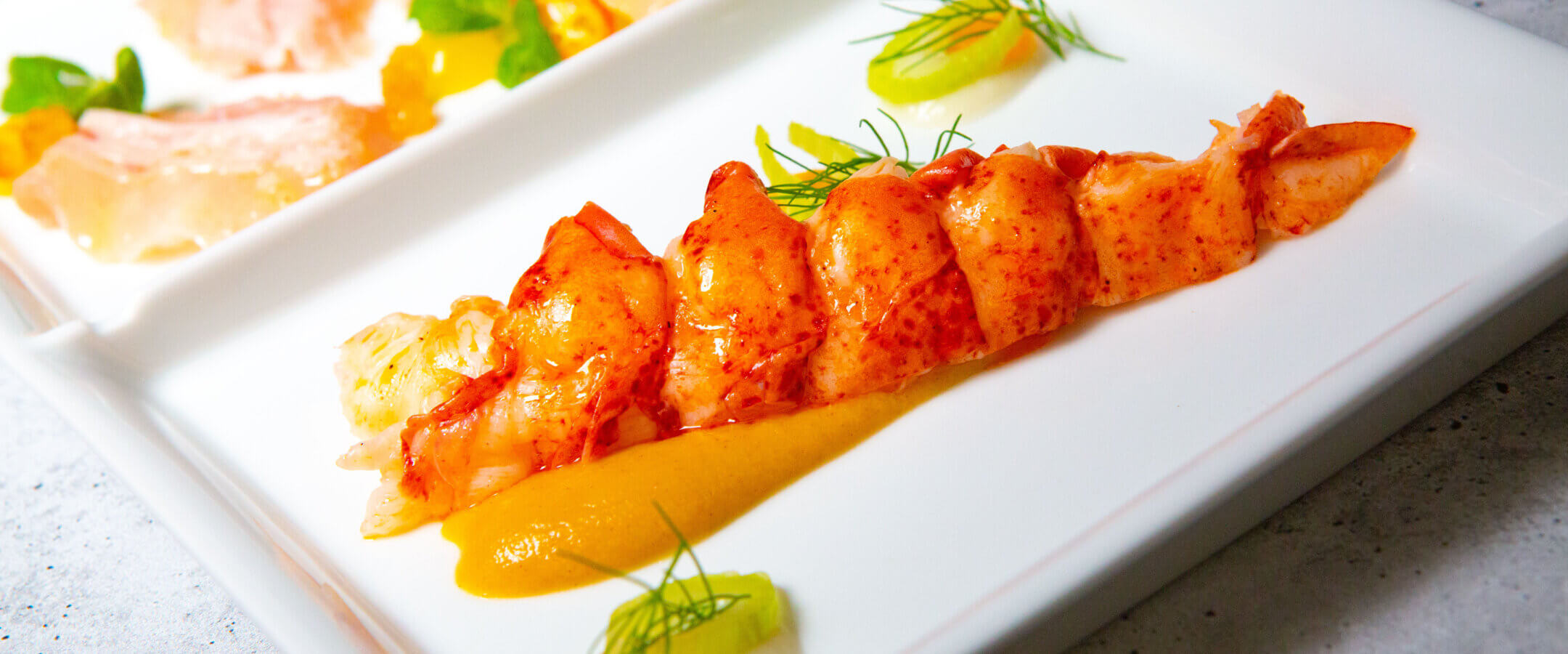 A neatly sliced lobster tail is plated with a smooth orange sauce, garnished with fresh herbs and small dollops of another sauce, on a white divided plate with additional seafood in the background.