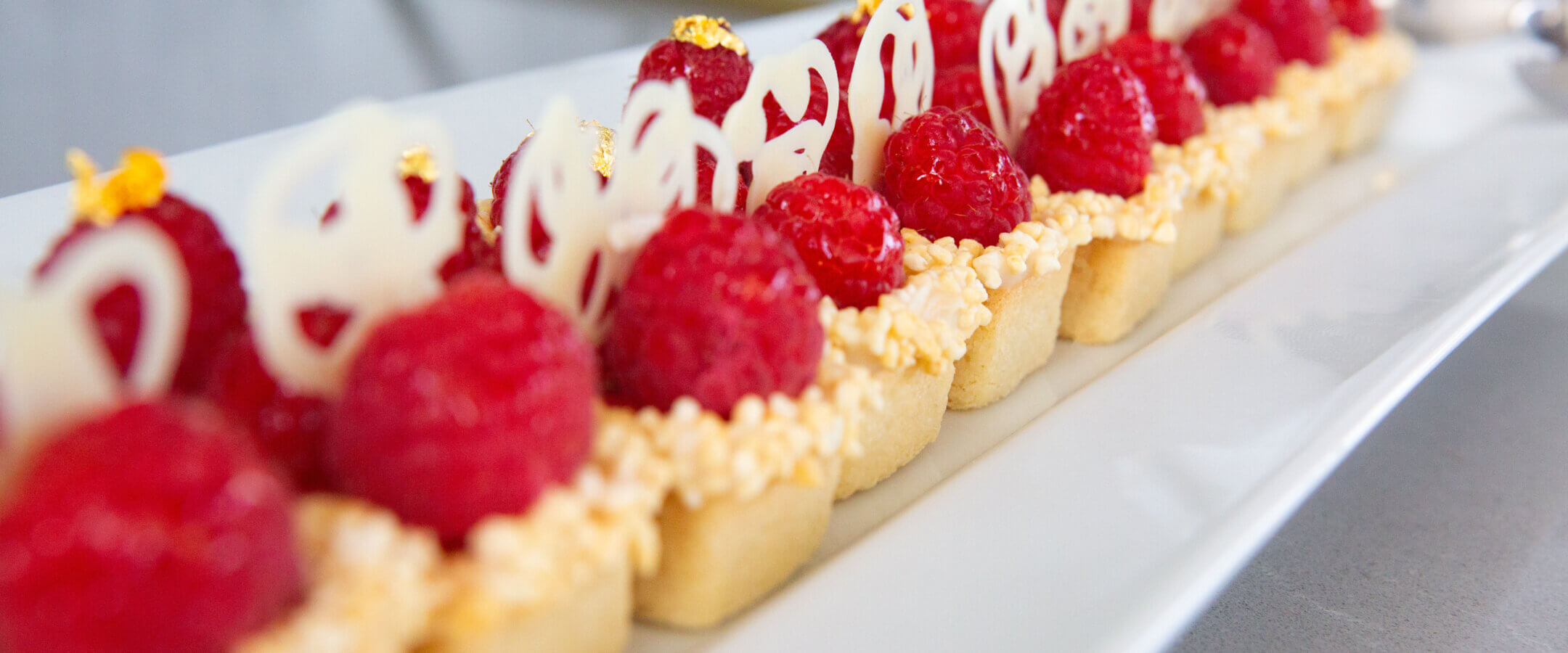 A row of elegant mini tart desserts topped with fresh raspberries, white chocolate decorations, and small gold leaf accents, arranged neatly on a rectangular white plate.
