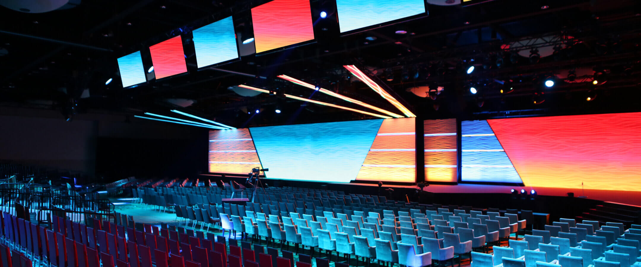 A large conference hall with rows of empty chairs facing a brightly lit stage and multiple colorful LED screens displaying abstract patterns in blue, red, and orange hues.