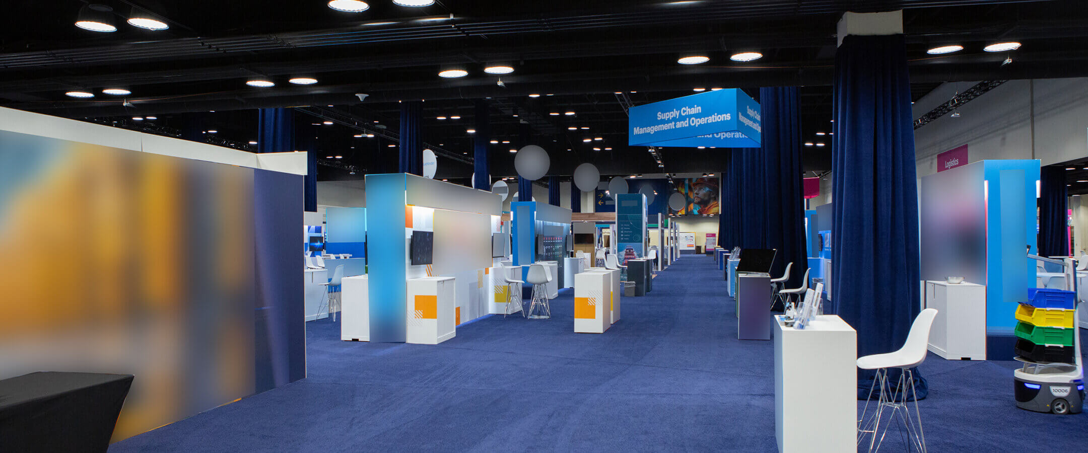 A spacious, modern exhibition hall with blue carpeting, white booths, display screens, chairs, and dividers. Overhead signs and ceiling lights are visible, creating a professional event or trade show atmosphere.