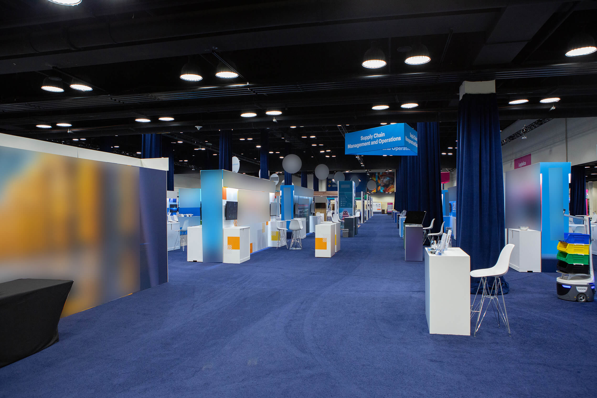 A spacious, modern exhibition hall with blue carpeting, white booths, display screens, chairs, and dividers. Overhead signs and ceiling lights are visible, creating a professional event or trade show atmosphere.