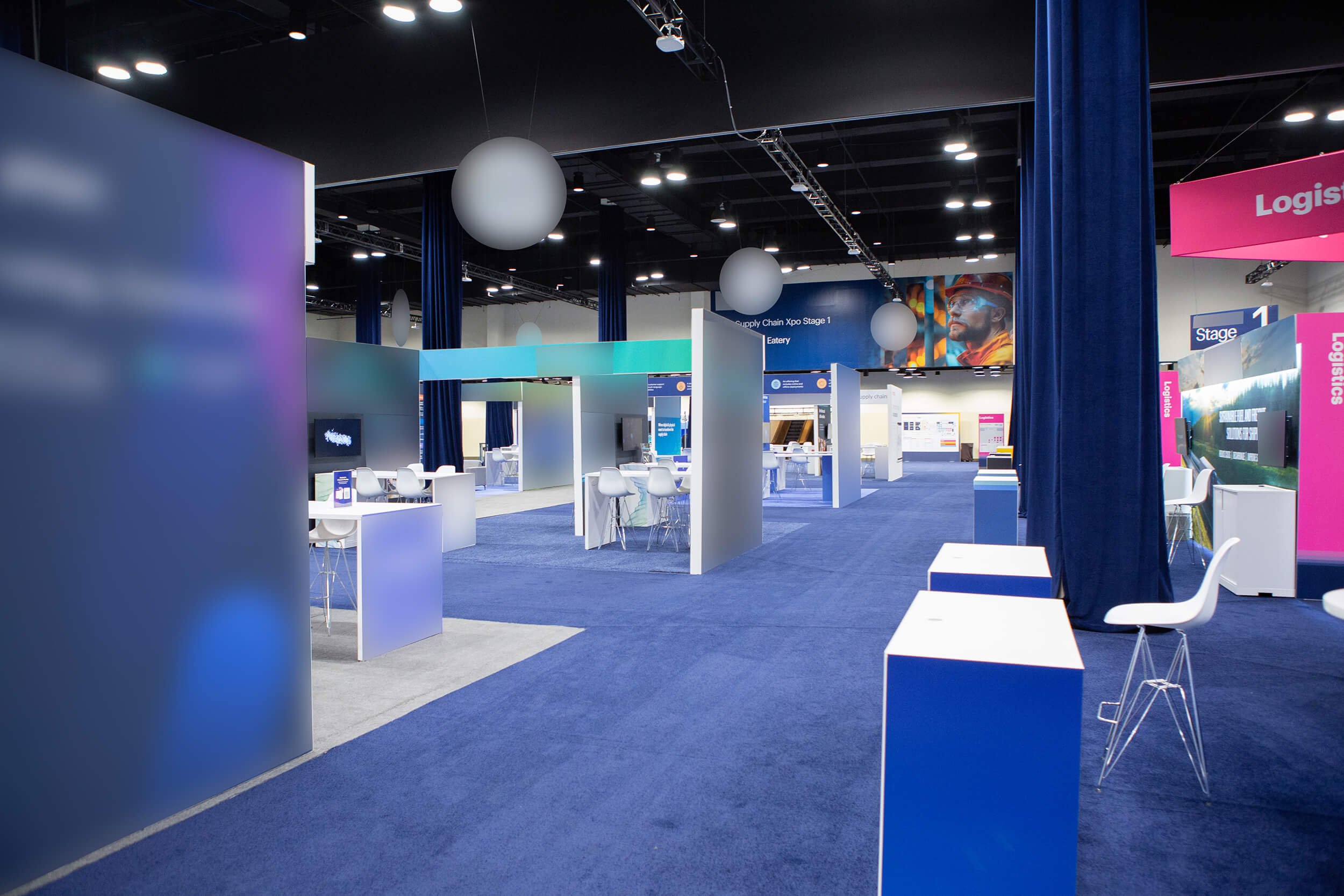 A modern convention center hall with blue carpet, partitioned booths, white tables and chairs, and colorful signs and banners, all brightly lit by overhead lights.