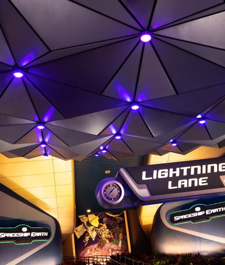 The entrance to the Spaceship Earth attraction at Epcot, featuring geometric panels above and illuminated signs that read “Lightning Lane” and “Spaceship Earth,” with purple lighting accentuating the structure.