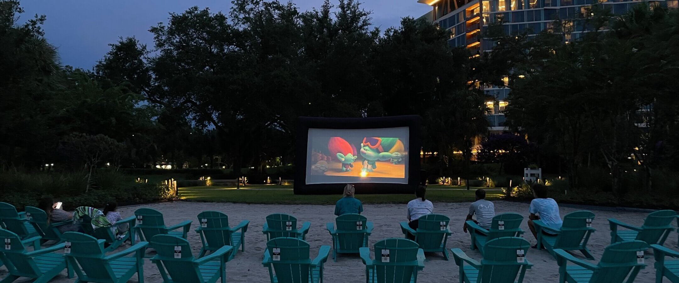 An outdoor movie is being shown on a large screen in front of rows of empty and sparsely occupied teal chairs on a sandy area at dusk, with a modern, well-lit building and trees in the background.