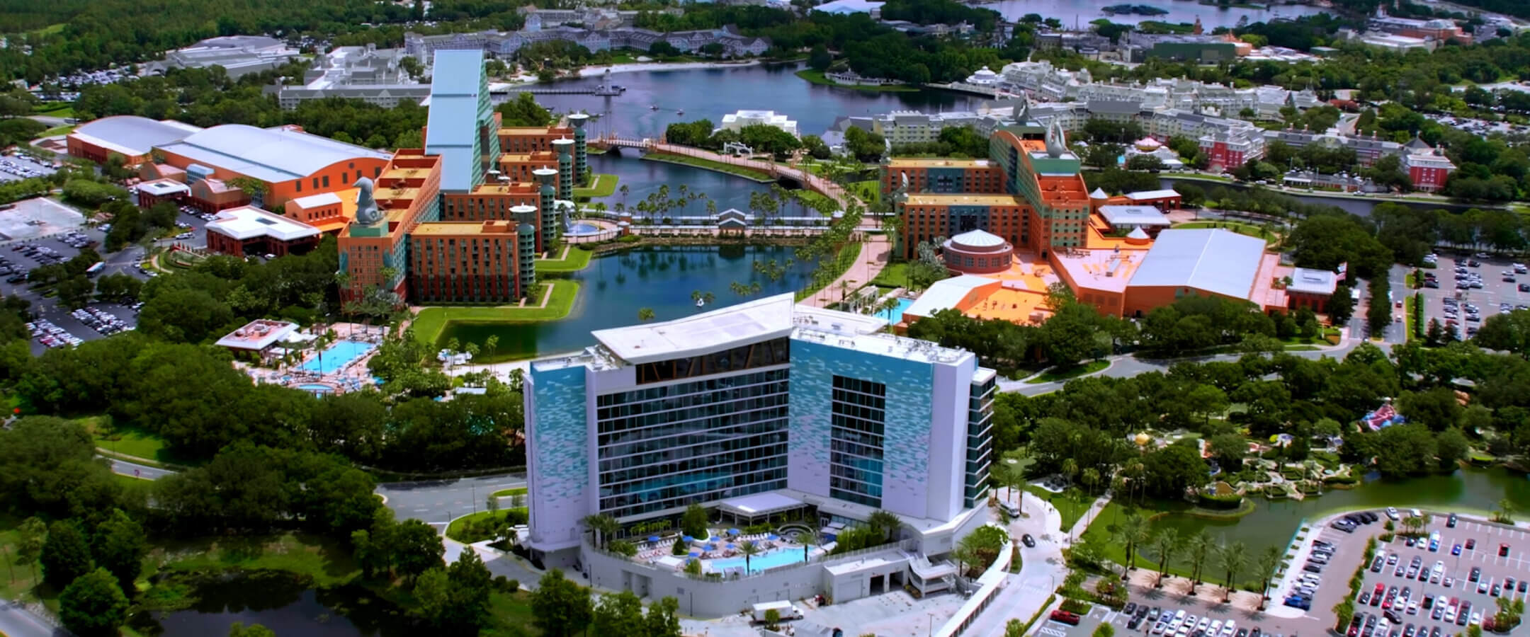 Aerial view of a hotel complex with modern and colorful buildings, surrounding pools, a lake, parking lots, and green spaces, with a theme park and geodesic dome visible in the background.