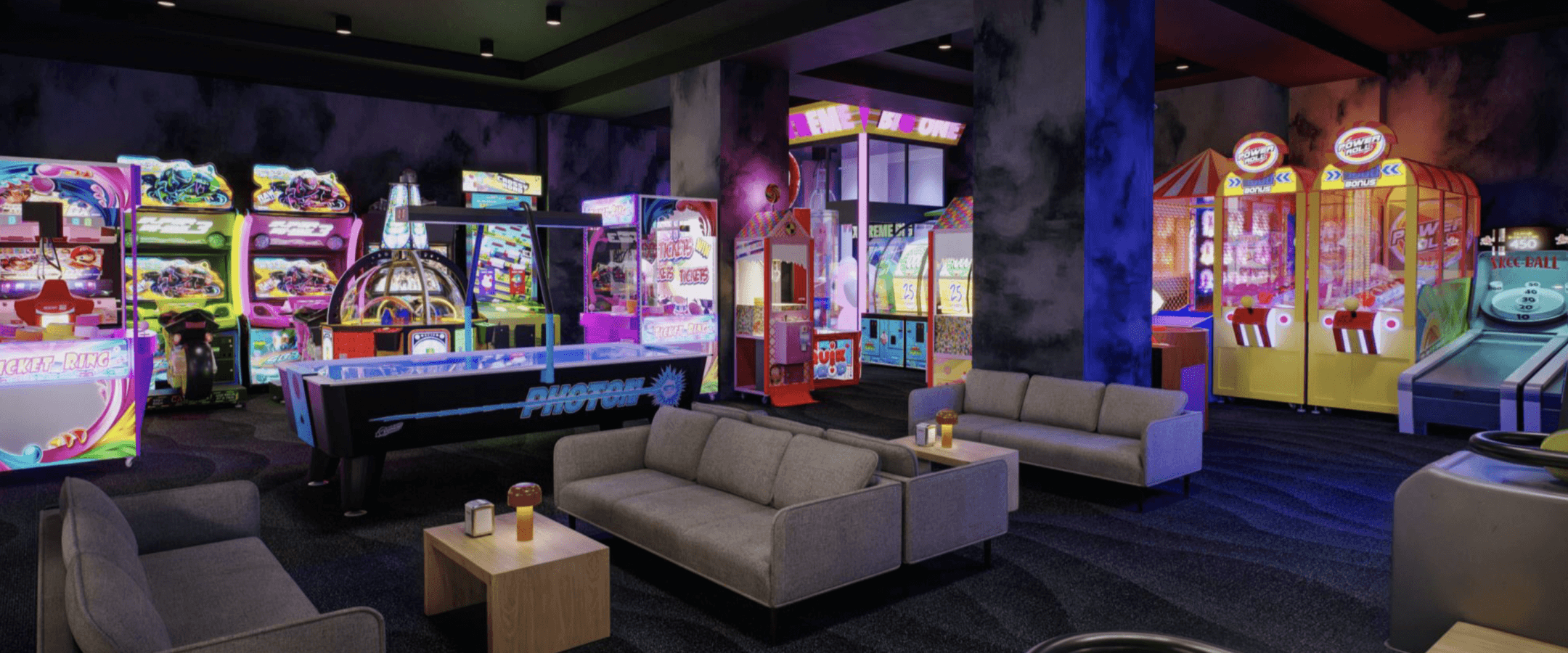 A modern arcade room with colorful game machines, air hockey, and skee-ball. Several gray couches and small wooden tables with drinks are arranged for seating in the foreground. The room is dimly lit.