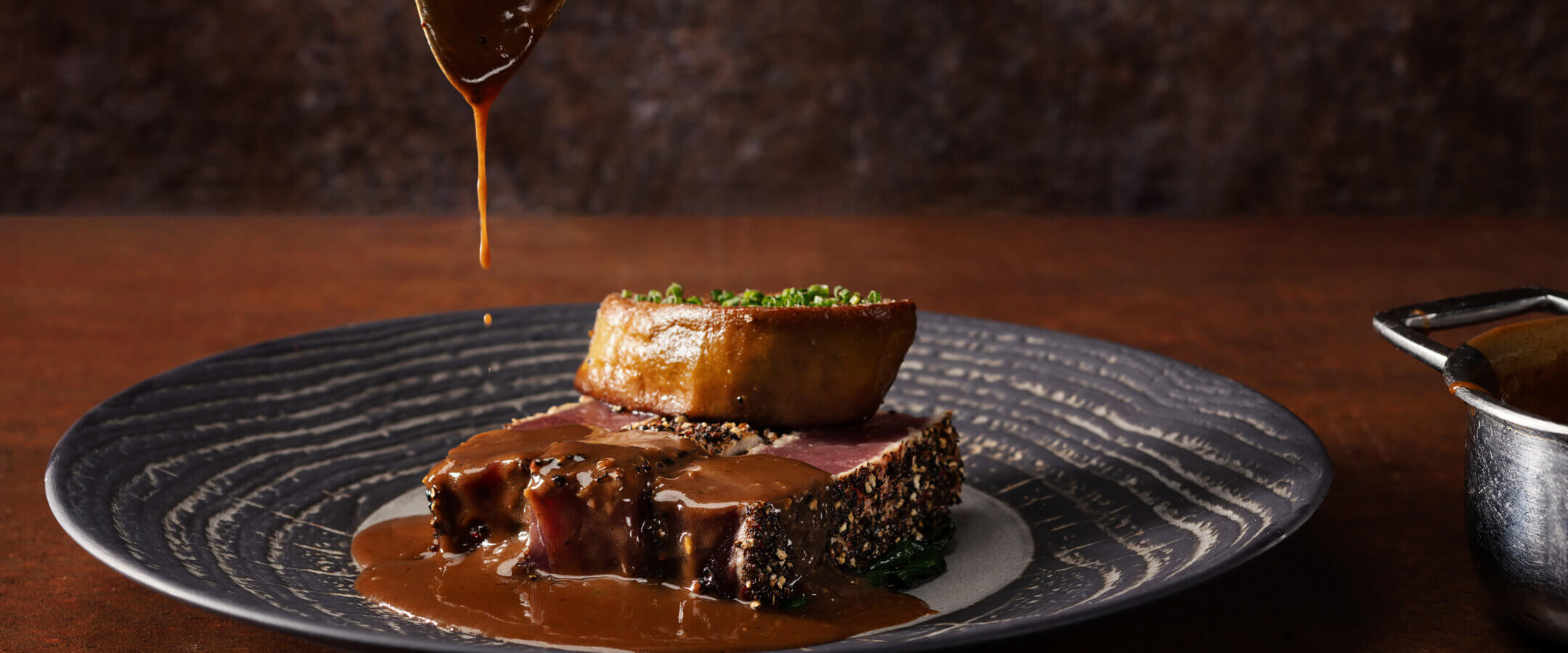 A close-up of a gourmet dish featuring a seared steak topped with a round piece of foie gras, served on a patterned plate, as rich brown sauce is being drizzled over the food from a spoon above.
