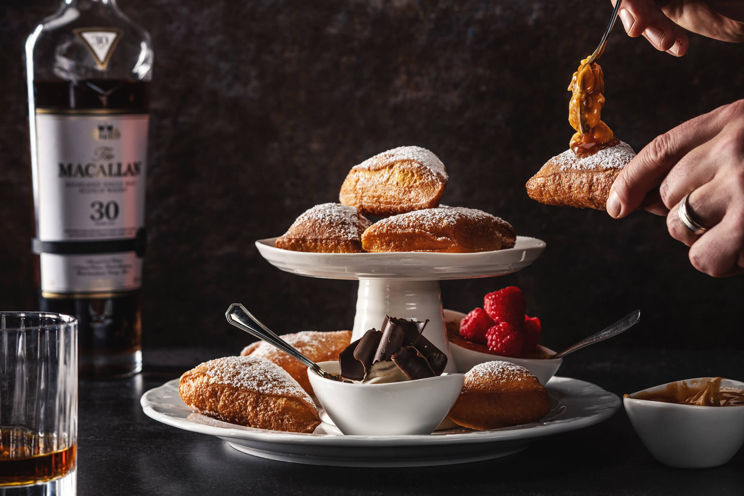 Eine Hand bestreicht einen Puderzuckerteig mit Karamell neben einem Ständer mit Beignets. Daneben liegen Himbeeren, Schokolade, Sahne, eine Flasche Macallan 30 Whisky und ein Glas Whisky auf einem dunklen Tisch.