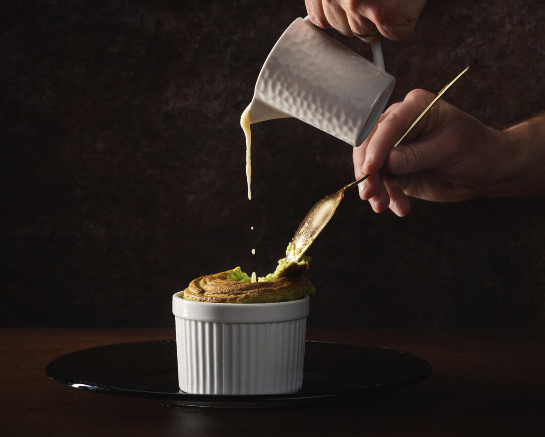 A hand holds a spoon breaking into a golden soufflé in a white ramekin while another hand pours creamy sauce over it from a small white pitcher against a dark background.