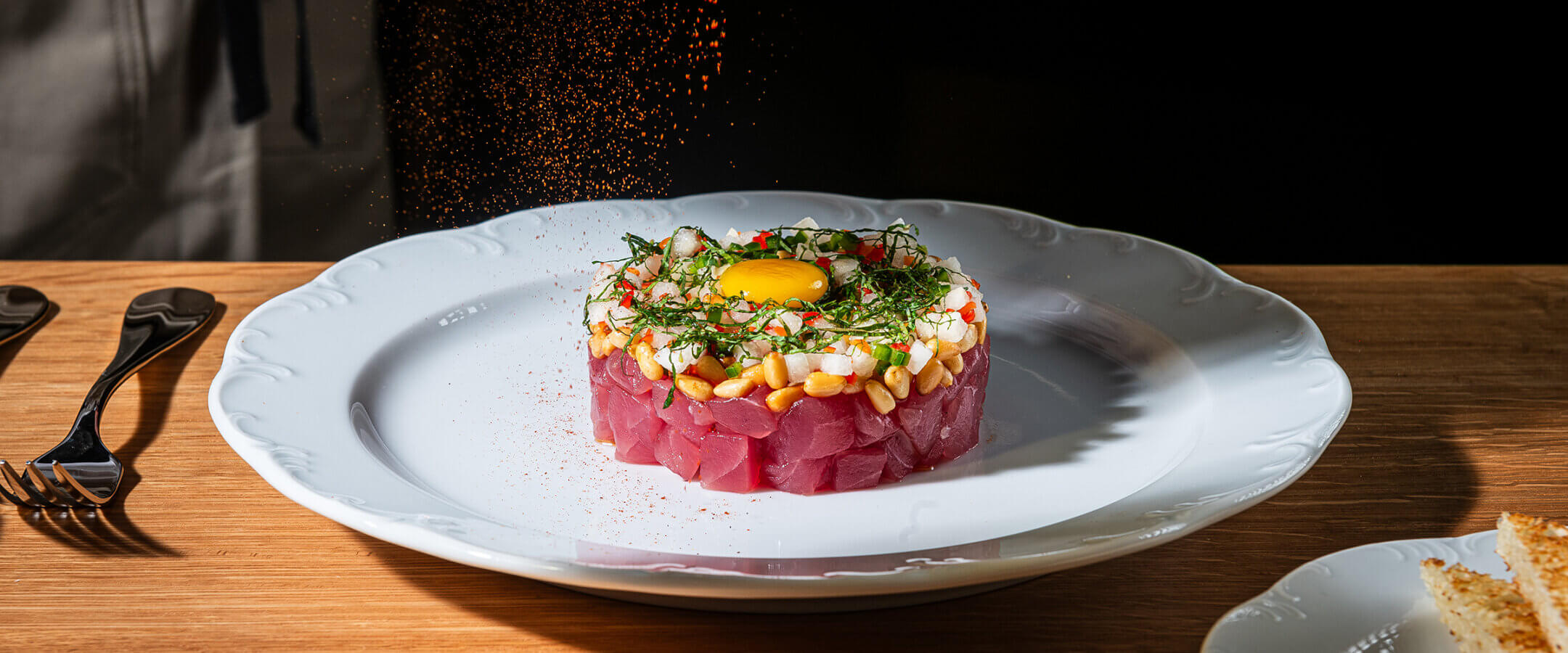 A hand sprinkles seasoning over a neatly plated tuna tartare topped with a raw egg yolk, herbs, and garnishes on a white plate, set on a wooden table with utensils and toast nearby.