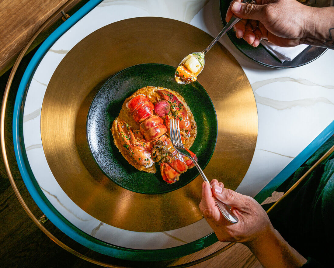 A person uses a fork and spoon to eat a gourmet dish featuring rolled seafood and sauce, elegantly plated on a dark green plate atop a gold charger on a marble-topped table.