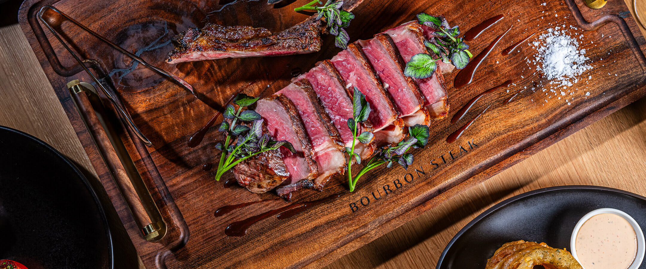 A sliced, medium-rare steak garnished with herbs is served on a wooden board with salt, a steak knife, and fork. Surrounding plates hold onion rings, dipping sauce, greens, and other side dishes.