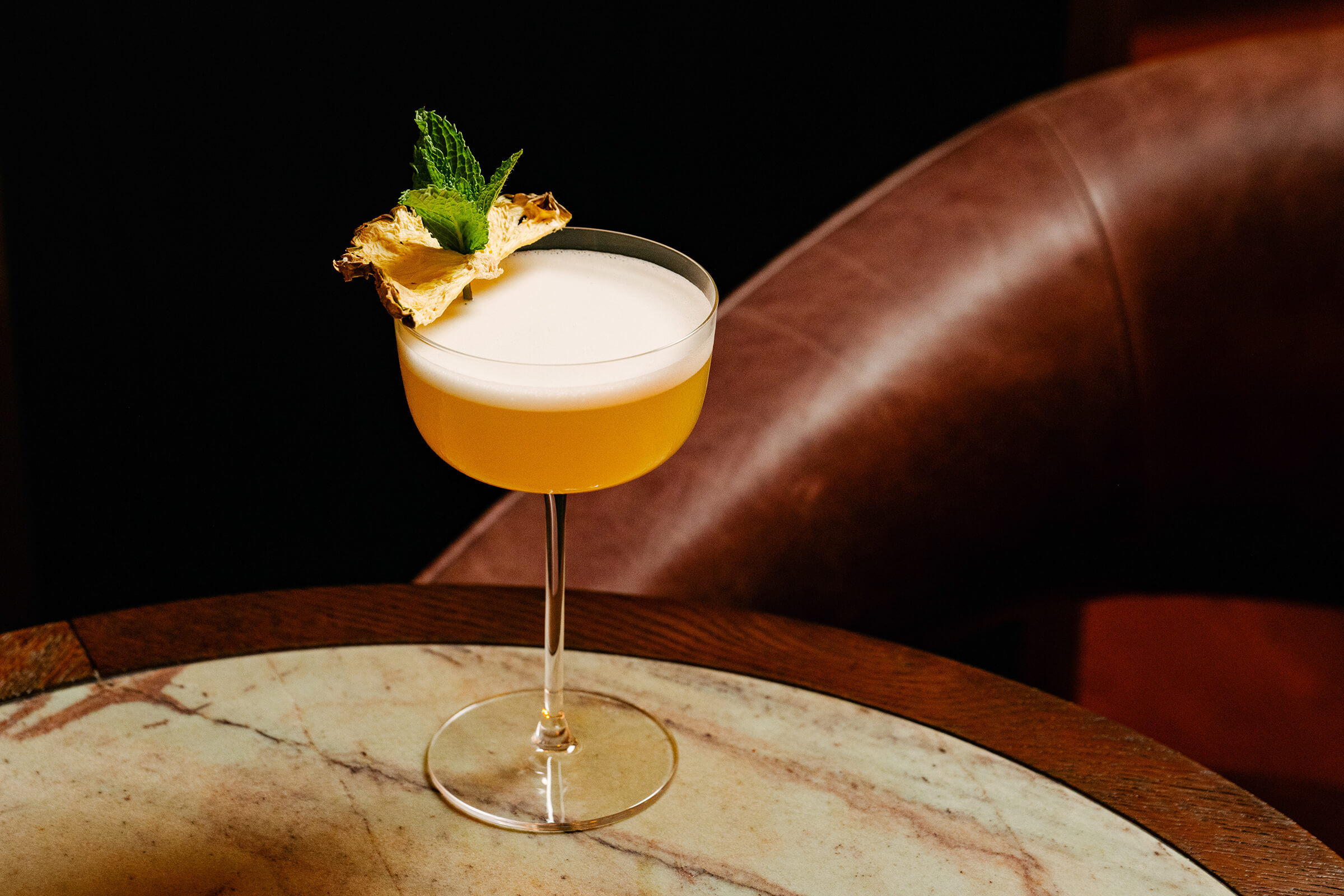 Ein gelber Cocktail mit Schaumkrone, serviert in einem Coupe-Glas, garniert mit einer getrockneten Ananasscheibe und einem Minzezweig, auf einem runden Marmortisch neben einem braunen Ledersessel.