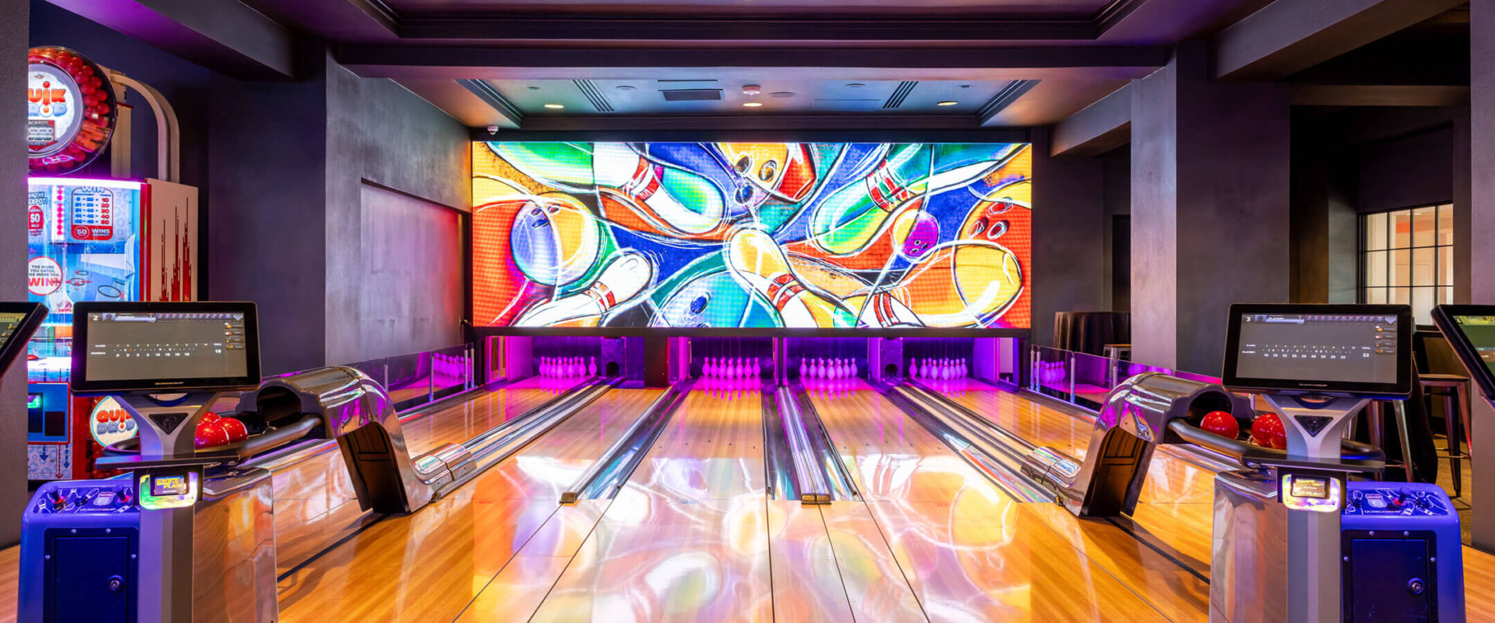 A modern bowling alley with four lanes, colorful bowling balls, electronic scoreboards, and a vibrant mural of bowling pins and balls on a large screen at the end of the lanes.