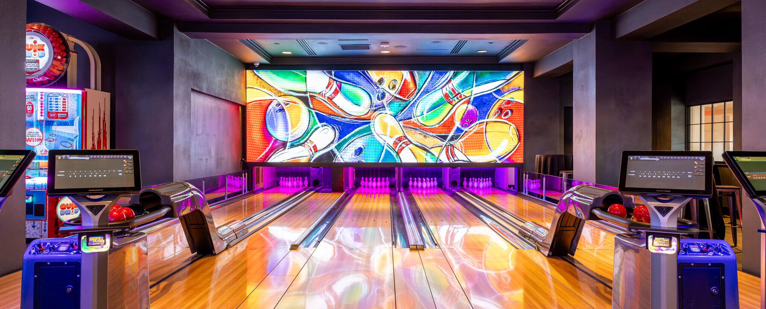A modern bowling alley with four lanes, colorful bowling balls, electronic scoreboards, and a vibrant mural of bowling pins and balls on a large screen at the end of the lanes.