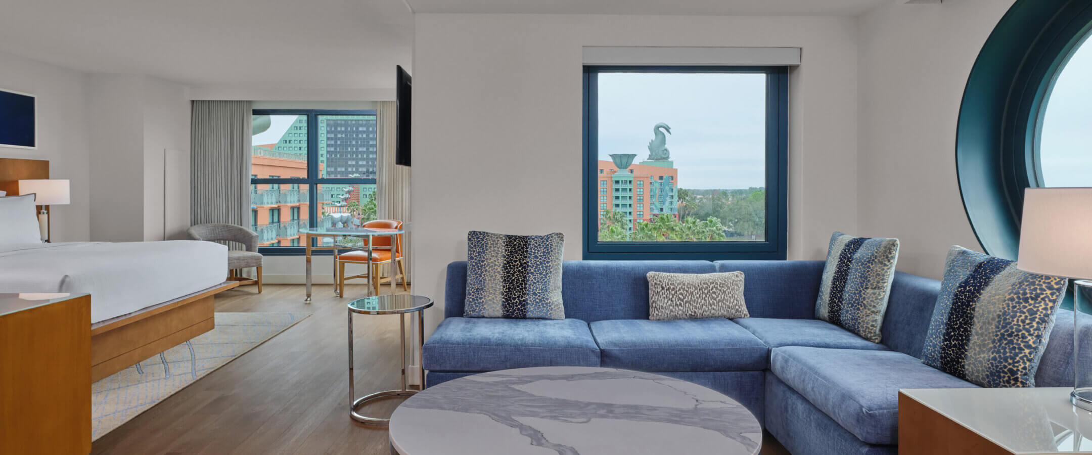 Modern hotel suite with a blue sectional sofa, round marble coffee table, and large windows offering a view of colorful buildings and a dolphin statue outside. A bed and desk are visible in the adjoining area.