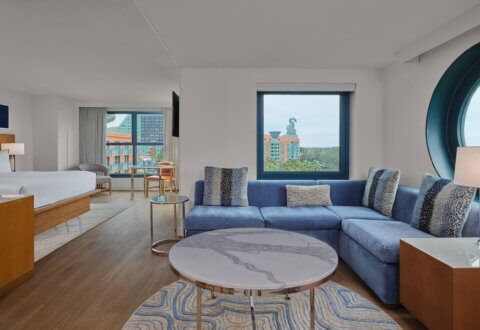Modern hotel suite with a blue sectional sofa, round marble coffee table, and large windows offering a view of colorful buildings and a dolphin statue outside. A bed and desk are visible in the adjoining area.
