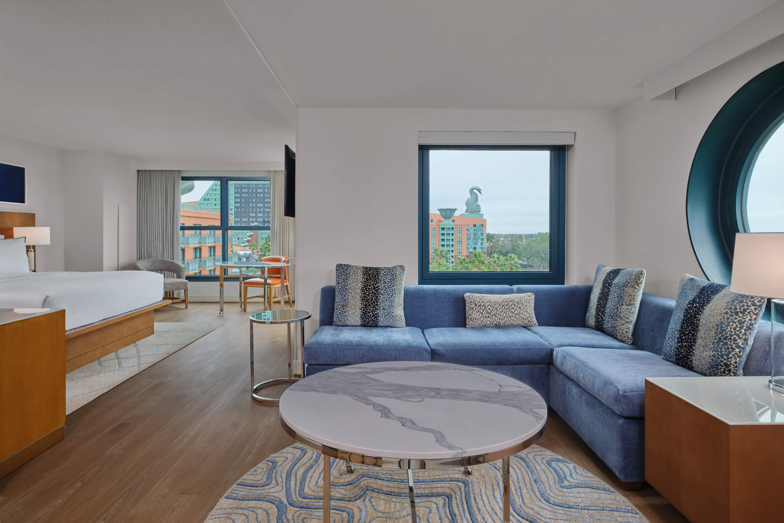 Modern hotel suite with a blue sectional sofa, round marble coffee table, and large windows offering a view of colorful buildings and a dolphin statue outside. A bed and desk are visible in the adjoining area.