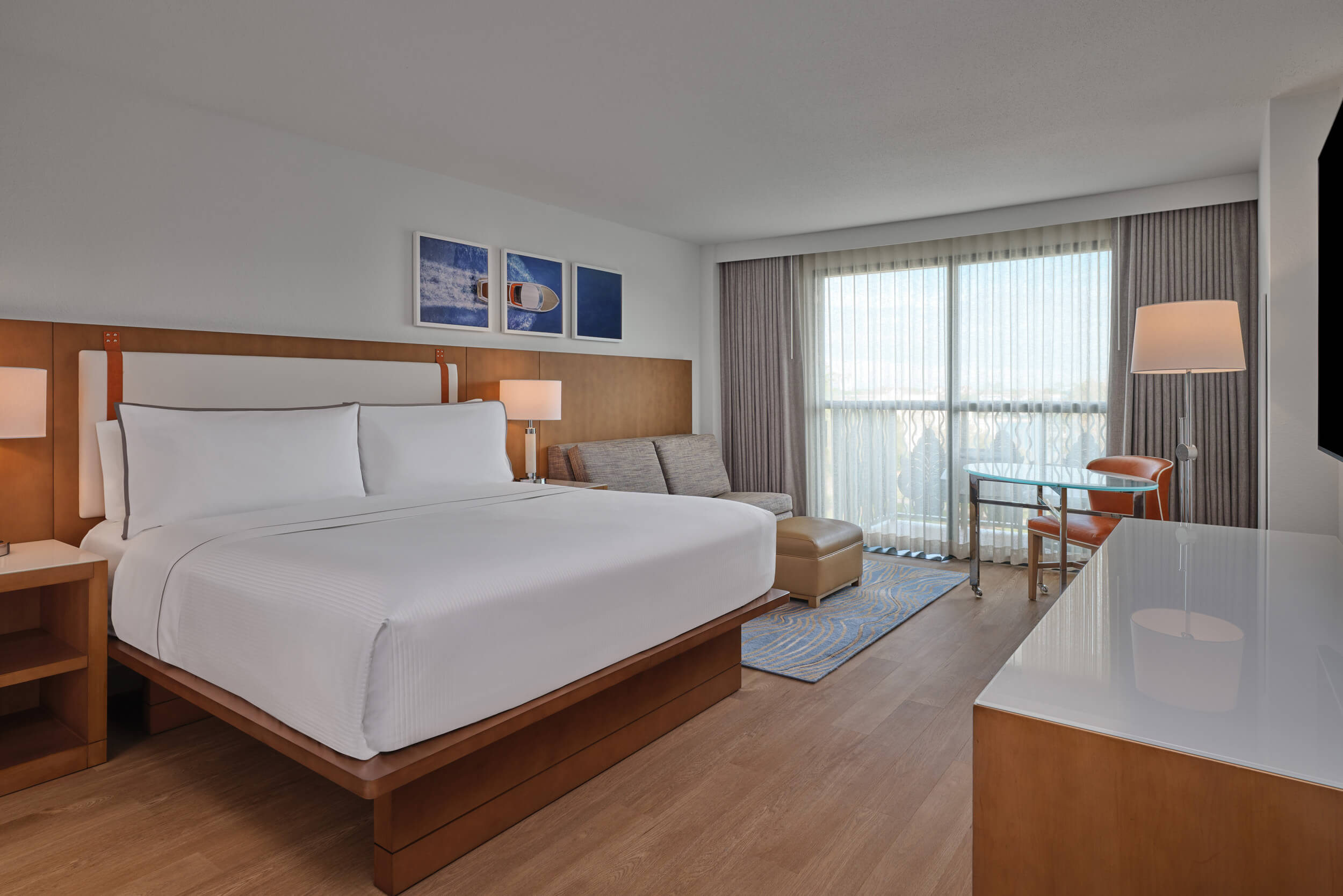 A modern hotel room with a large bed, a gray sofa, a small table with a chair by the window, a floor lamp, and minimalistic decor. Light wood furniture and large windows let in natural light.