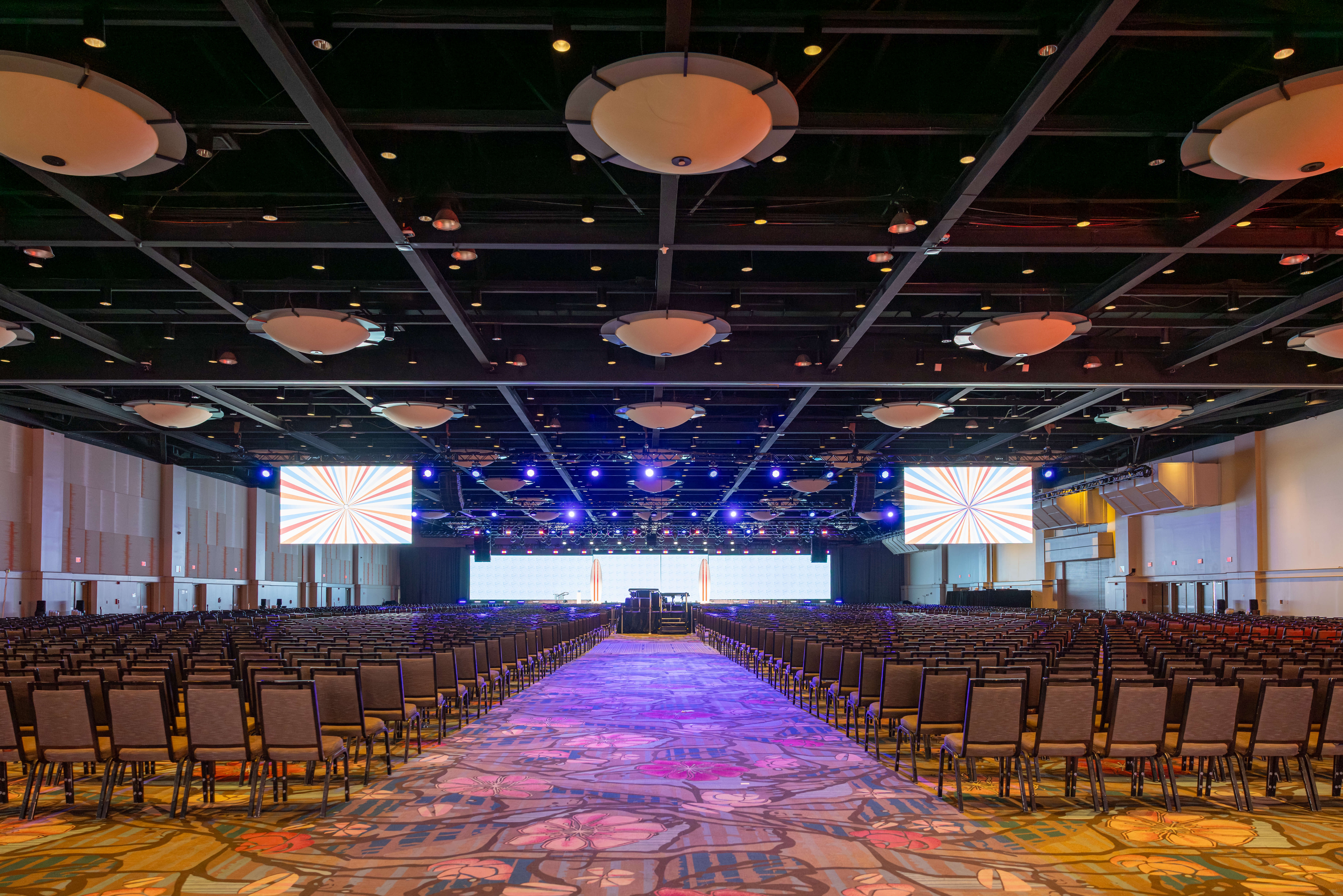A large, empty conference hall with rows of chairs facing a stage, large projection screens, and colorful lighting on the ceiling above a patterned carpeted floor.