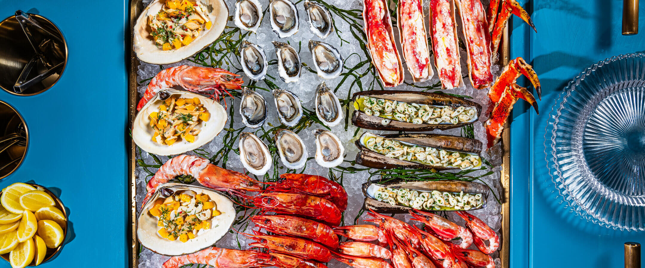 A seafood platter on ice features oysters, crab legs, large shrimp, scallops in shells, and stuffed zucchini halves, with lemon wedges on the side, all arranged on a blue tray.