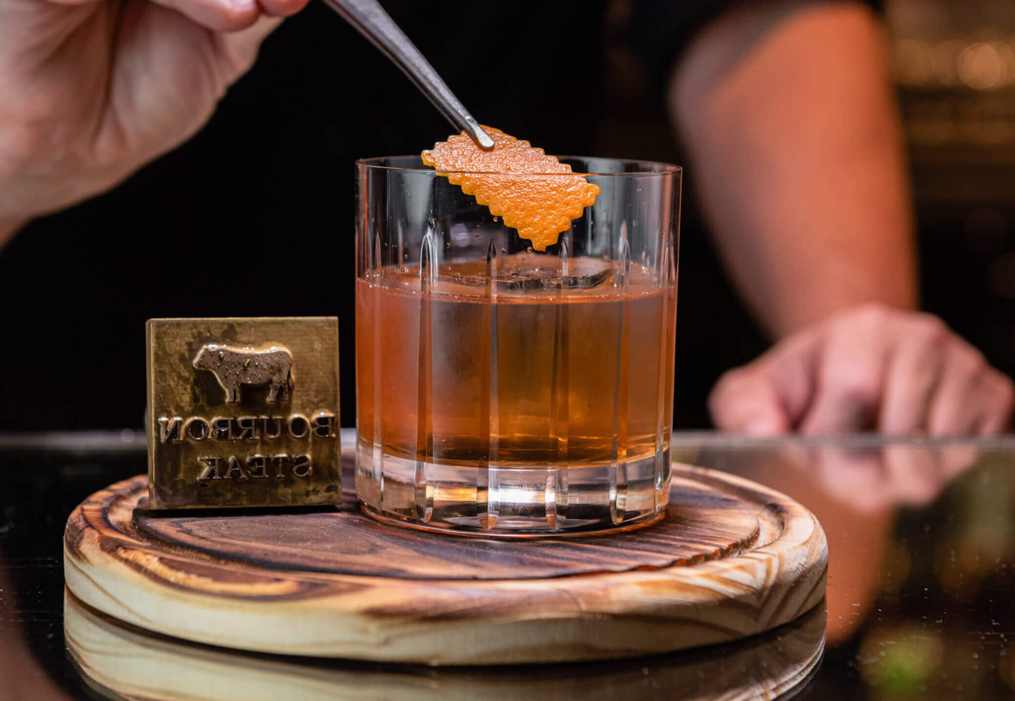Eine Hand benutzt eine Pinzette, um eine Orangenschale auf einem Cocktail in einem Glas mit Eis zu platzieren, das auf einem hölzernen Untersetzer neben einer Bourbon Steak steht.