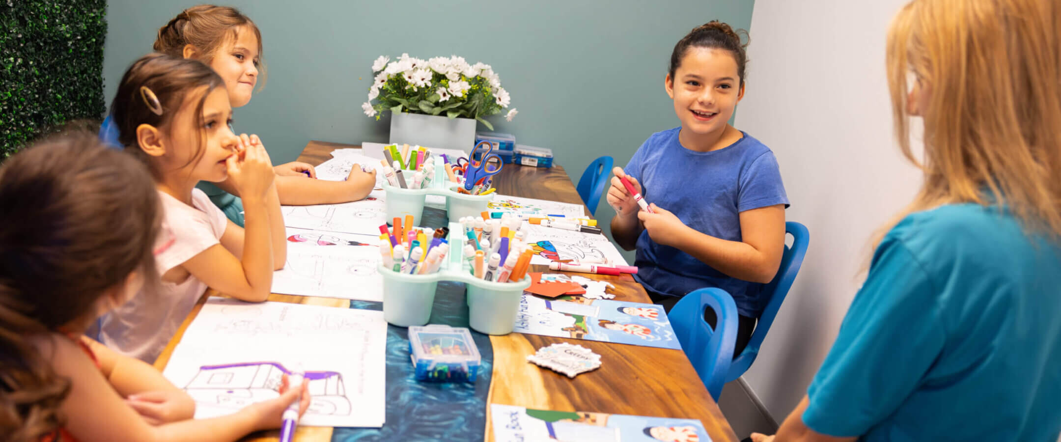Four young girls sit around a table doing arts and crafts with markers and papers, while a female adult sits with them, smiling in a bright, cheerful classroom.