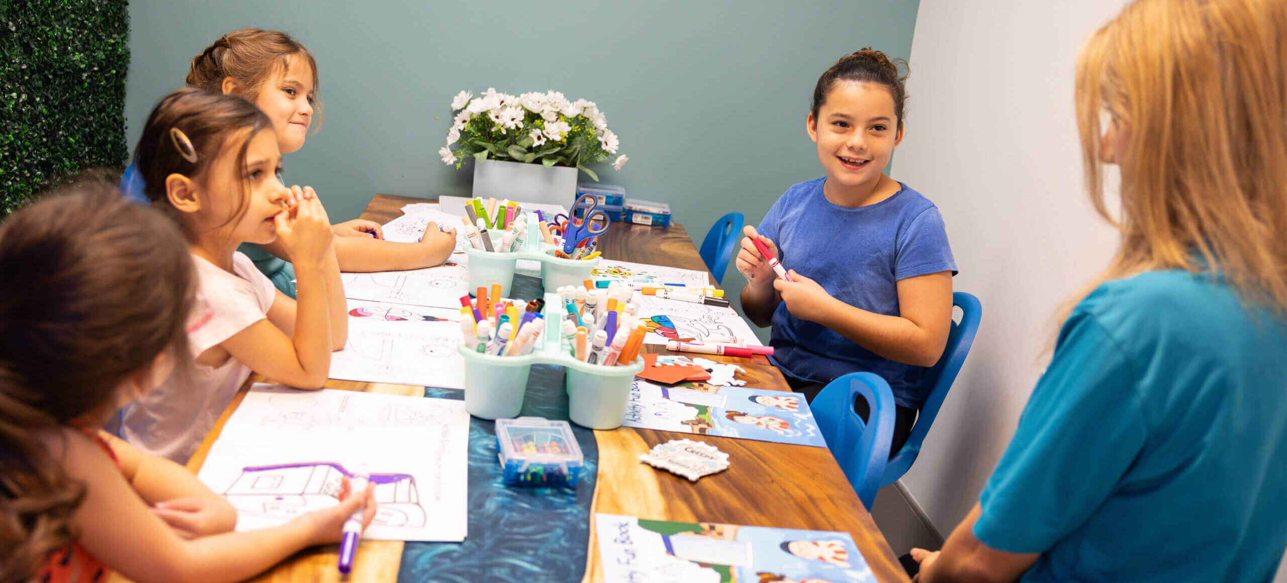 Four young girls sit around a table doing arts and crafts with markers and papers, while a female adult sits with them, smiling in a bright, cheerful classroom.