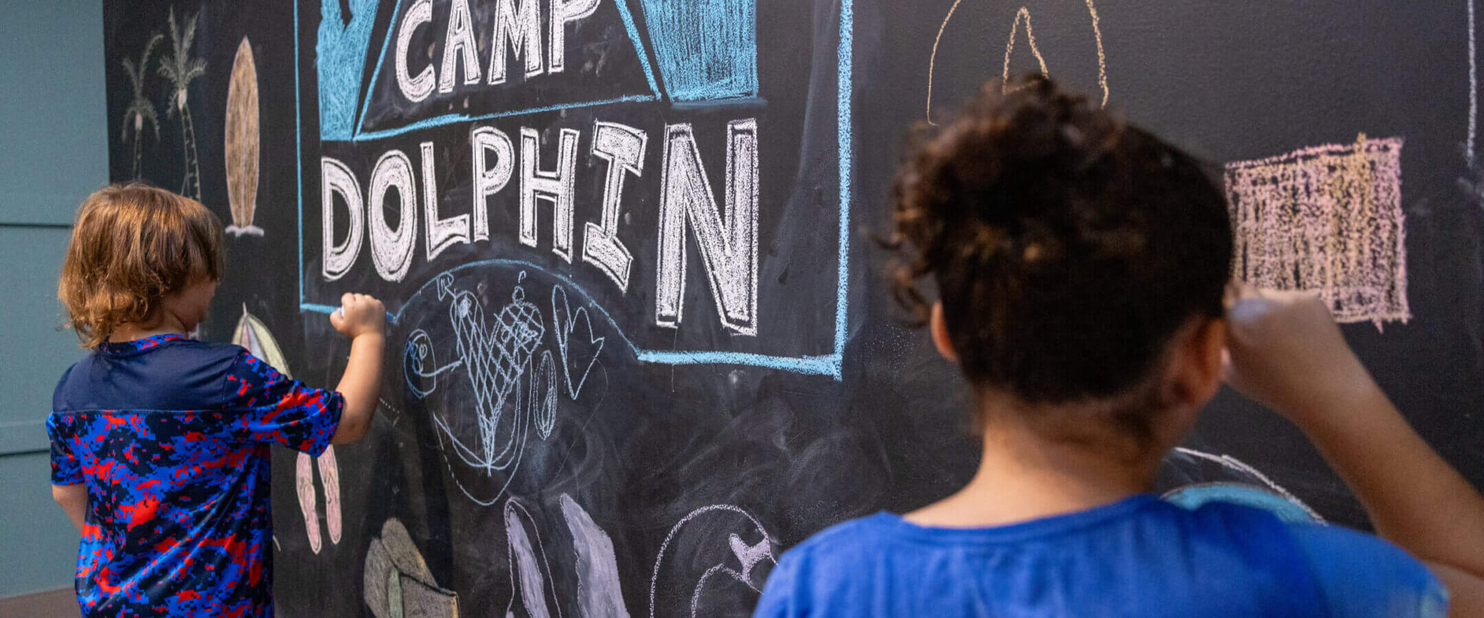 Two children are drawing colorful pictures and the words CAMP DOLPHIN with chalk on a large black chalkboard wall. The scene is creative and playful.