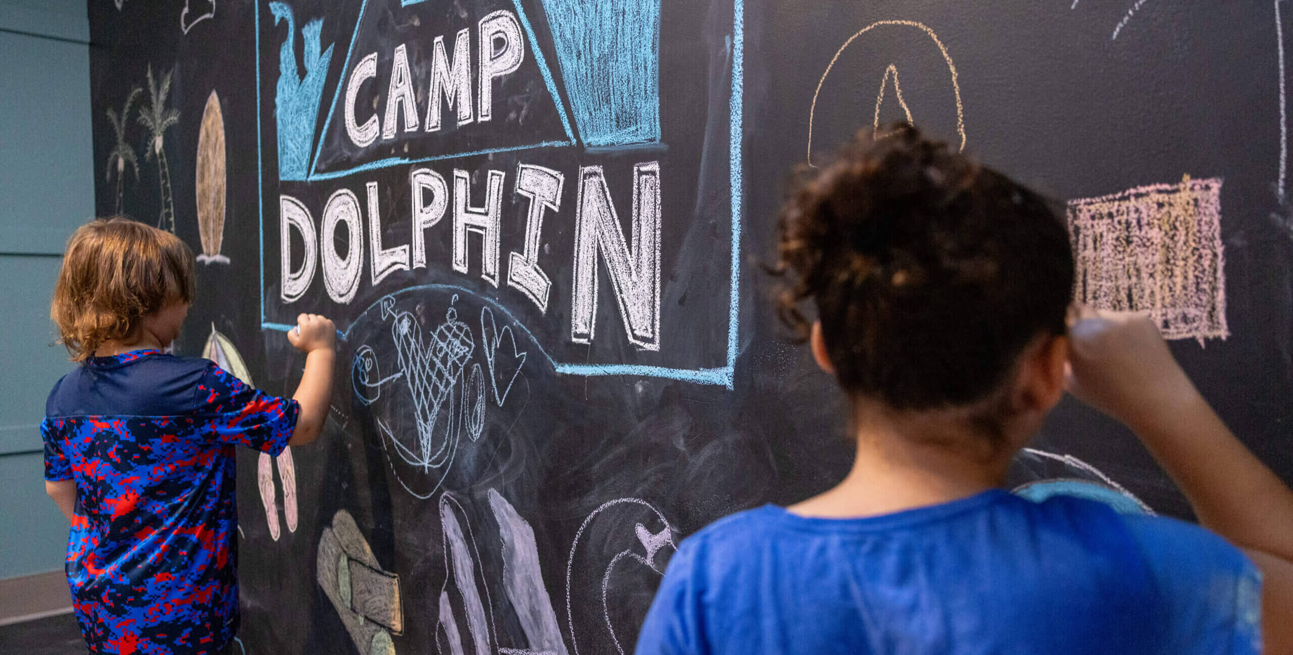 Two children are drawing colorful pictures and the words CAMP DOLPHIN with chalk on a large black chalkboard wall. The scene is creative and playful.
